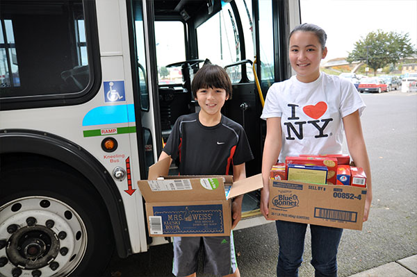 two young people stand outside a city bus holding boxes of donated food