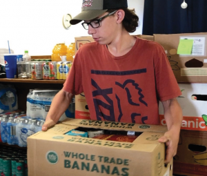 a person in a baseball cap glasses and red t-shirt moves a banana box full of food