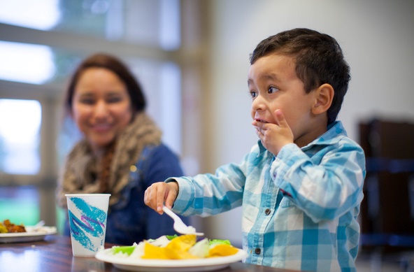 child with short dark hair eats food with fork and hand while mom looks on