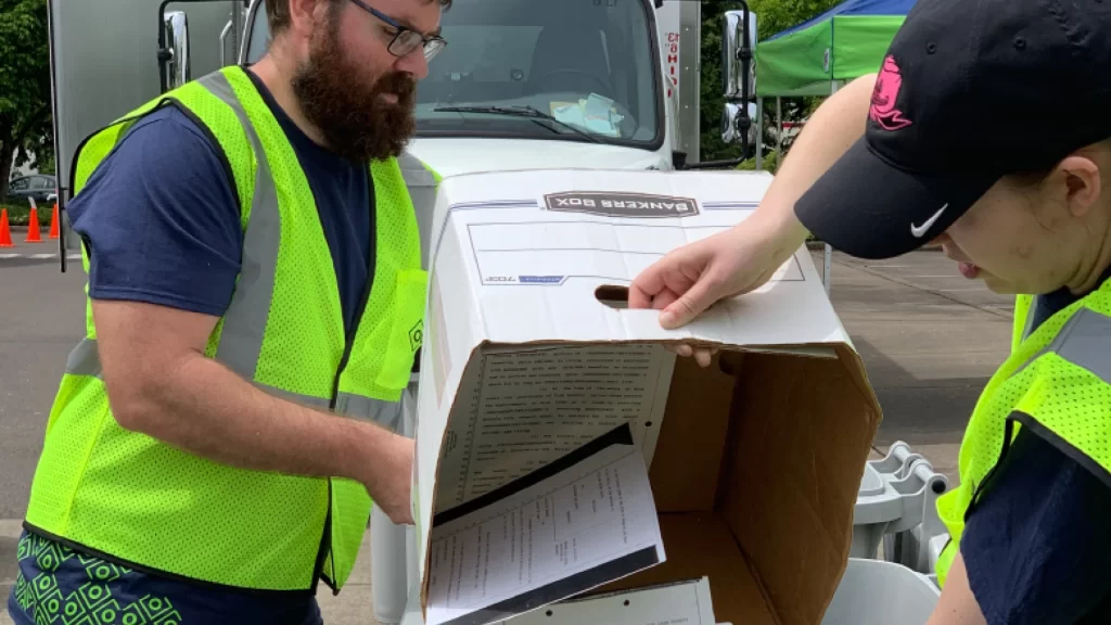 two people in neon green vests dump a box of papers into another receptacle
