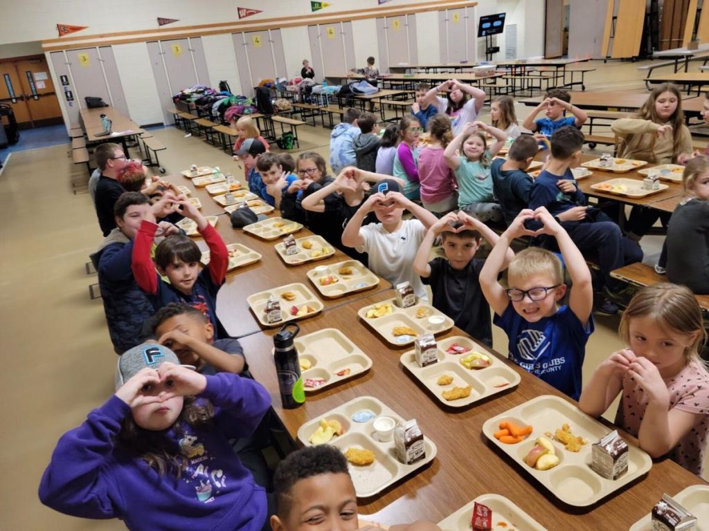 kids sit at table with lunch trays making the O symbol with their hands